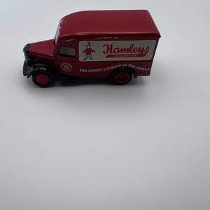 May include: A red toy delivery truck with black wheels. The truck has the Hamleys logo on the side, with the text "THE FINEST TOYSHOP IN THE WORLD". The truck is a vintage style toy.