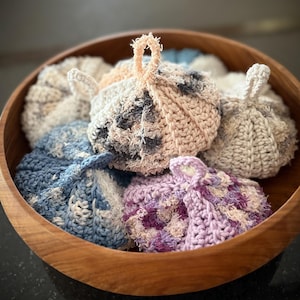 May include: A collection of round, crocheted scrubbers with small hanging loops, featuring textured yarn in various colours including blue, purple, cream, and grey, all contained within a smooth, circular wooden bowl.