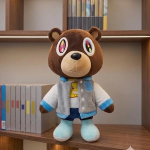 May include: A brown plush bear toy wearing a grey and white varsity jacket with blue trim, a white t-shirt featuring a yellow graphic, and blue shorts. The bear has large eyes with colorful, multi-segmented pupils and a black nose.