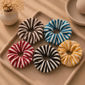 May include: A set of five circular hair scrunchies made of knit fabric, each featuring a distinct two-tone striped pattern. The colors include brown and white, blue and white, red and white, black and white, and yellow and white, arranged in a circular formation.