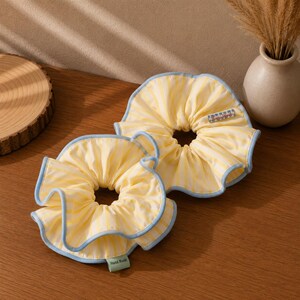 May include: Two fabric hair scrunchies featuring a yellow and white vertical striped pattern with light blue ruffled edges. One scrunchie displays a small rectangular tag with text, while the other features a green tag labeled Hand Made.