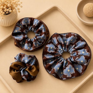 May include: Three hair scrunchies featuring a dark brown fabric base with a repeating pattern of light blue cloud-like shapes and small yellow star icons. The scrunchies vary in size and fullness, showcasing a gathered, ruffled texture throughout.