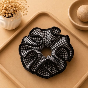 May include: A hair scrunchie featuring a black and white gingham check pattern. The fabric is gathered into a circular, ruffled shape with a solid black fabric trim along the outer and inner edges, creating a distinct contrast against the checkered print.