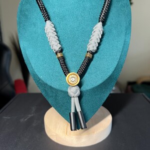 Può includere: Una collana con cordino intrecciato nero, due sezioni in paracord grigio, distanziatori dorati con motivo greco e una base centrale a forma di bossolo dorato con testo. Il cordino termina con un nodo grigio e due punte nere.