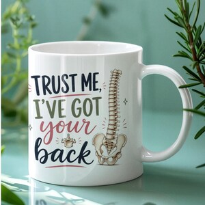 May include: White ceramic mug with the text "TRUST ME, I'VE GOT your back" in black, green, and pink. A spine and pelvis illustration is on the right side of the mug. The mug has a white handle.