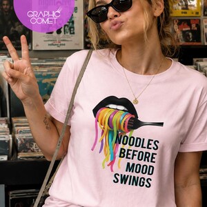 Può includere: Una t-shirt rosa chiaro con una grafica di labbra nere che tengono una forchetta nera con spaghetti arcobaleno colorati. Sotto la grafica, il testo recita NOODLES BEFORE MOOD SWINGS in grassetto nero. La t-shirt ha maniche corte e girocollo.