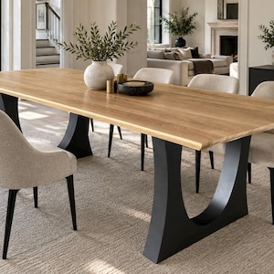 Puede incluir: Una mesa de comedor rectangular con una superficie de madera clara y dos bases de pedestal negras en forma de U. La mesa está rodeada por seis sillas tapizadas con tela color crema y patas negras. Un jarrón blanco con vegetación está sobre la mesa.