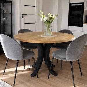 Peut inclure: Une table à manger ronde en bois avec une finition brun clair naturel et une base en métal noir incurvée, accompagnée de quatre chaises rembourrées grises avec des pieds fins en métal noir. Un vase en verre transparent avec des fleurs blanches.