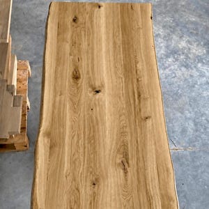 Pode incluir: Um tampo de mesa retangular de madeira castanho-clara com uma borda natural em ambos os lados. A superfície exibe um padrão de grão de madeira proeminente com nós visíveis e variações orgânicas na textura da madeira.