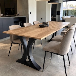 Peut inclure: Une table à manger rectangulaire avec un plateau en bois clair et deux pieds centraux en métal noir incurvés. Six chaises avec un revêtement en tissu beige et des pieds fins en métal noir sont disposées autour de la table, portant un bol sombre.