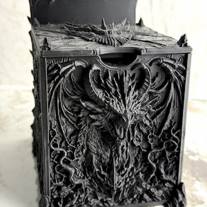 May include: A matte black square storage box featuring a highly detailed, three-dimensional relief of a dragon on the front panel. The dragon has large horns, spread wings, and intricate scale textures. The lid has a matching dragon wing design.