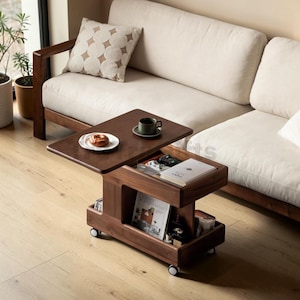 May include: A dark brown wooden coffee table on wheels with a lift-top surface. The table features an open storage compartment containing a book, a game controller, and various items, with a lower shelf holding magazines and small containers.