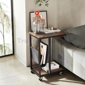 May include: A rectangular side table with a dark metal frame and two wood-grain shelves. It features four caster wheels, a vertical fabric magazine holder, and a top surface holding a glass vase with flowers and two stacked books.