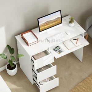 May include: A white computer desk featuring a rectangular top, a pull-out keyboard tray, and three stacked drawers on the left side. The desk surface holds a computer monitor, a white keyboard, a mouse, a brown document tray, and a notebook.