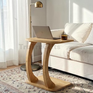 May include: A C-shaped side table crafted from light-toned wood with a smooth, curved support base. The table features a rectangular top surface that holds a silver laptop and a glass mug filled with a dark beverage, providing a functional workspace.