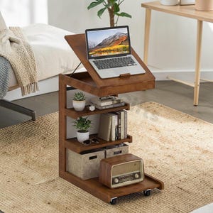 May include: A brown wooden laptop desk with multiple shelves. A laptop sits on the top surface, angled upwards. The shelves hold books, plants, and a vintage radio. The desk is on wheels for easy movement.