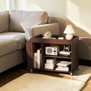 May include: A dark wood rolling side table with multiple storage shelves, featuring a top surface holding a round clock, a small lamp, and a cup. The shelves contain books, a vintage-style radio, white boxes, and a framed photograph.