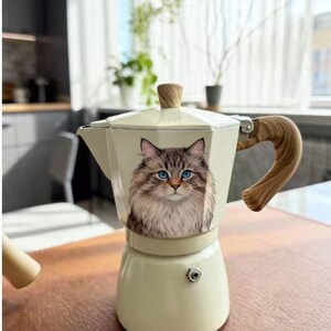 May include: Cream-colored stovetop espresso maker with a cat portrait. The cat has blue eyes and brown and white fur. The handle and lid knob are made of wood. The espresso maker is sitting on a brown wooden table.