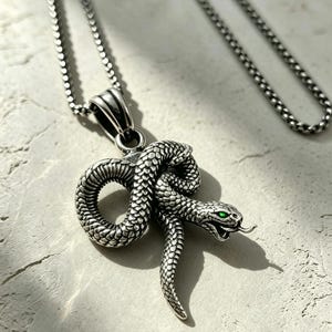 May include: A silver-toned metal necklace featuring a coiled snake pendant with detailed scales and a protruding tongue. The snake has a single vibrant green eye visible on the side of its head. The pendant hangs from a matching silver-toned chain.