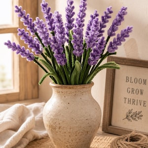 May include: A ceramic vase holds a bouquet of lavender-colored artificial flowers with green stems. The vase is beige and sits on a burlap surface. A wooden sign with the words "BLOOM GROW THRIVE" is in the background.