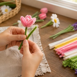 May include: A pink pipe cleaner tulip flower with a green stem, surrounded by various pipe cleaner flowers including daisies and lavender, and bundles of colorful pipe cleaner sticks in white, yellow, pink, green, and purple hues.