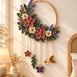 May include: A circular wall hanging featuring a wooden hoop adorned with macrame flowers in pink, white, and yellow, green leaves, and purple sprigs. Five cords hang from the bottom, ending in a small flower, a leaf sprig, a butterfly, a flower, and a moon.