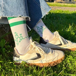 May include: A pair of beige sneakers with black accents and brown soles are shown on a grassy surface. The sneakers are paired with white socks featuring a green stripe and a chemical structure design. The person is wearing blue denim jeans.