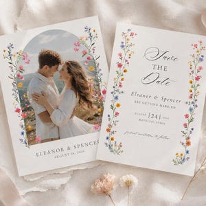 Pode incluir: Dois cartões retangulares de Save the Date com uma borda floral de flores silvestres coloridas. Um cartão exibe uma foto de um casal em uma moldura arqueada com o texto Eleanor & Spencer August 24, 2026. O outro cartão tem o texto Save the Date.