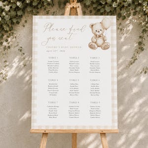 Puede incluir: Un cartel rectangular de distribución de asientos con un borde de cuadros beige y blanco. El texto dice Please find your seat, Phoebe's Baby Shower, April 22nd, 2024, y enumera las mesas del uno al nueve. Se ilustra un oso de peluche marrón con un globo.