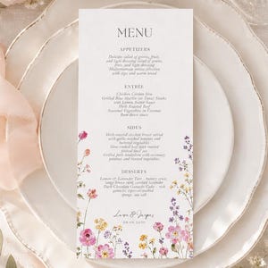 Pode incluir: Um cartão de menu branco vertical com uma borda floral em aquarela na parte inferior, apresentando flores silvestres rosas, amarelas e roxas. O texto inclui títulos para entradas, pratos principais, acompanhamentos e sobremesas, com os nomes Laura e Jasper.