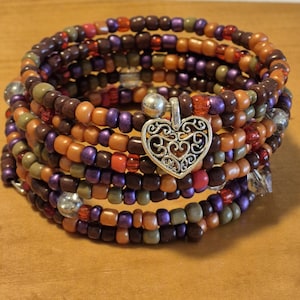 May include: A multi-strand wrap bracelet composed of small glass beads in shades of brown, orange, purple, and olive green. The bracelet features a silver-toned metal heart charm with an intricate filigree cutout design attached to the center.