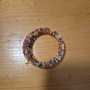 May include: A multi-strand beaded bracelet with a spiral design. The beads are a mix of colors, including red, yellow, purple, silver, and cream. The bracelet is coiled and rests on a wooden surface.