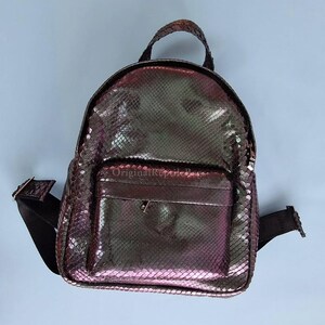 Puede incluir: Una mochila pequeña con un acabado texturizado de piel de serpiente iridiscente en tonos púrpura y verde oscuro. Cuenta con un asa superior, un bolsillo frontal con cremallera con el texto OriginalReptileBags y correas negras ajustables.