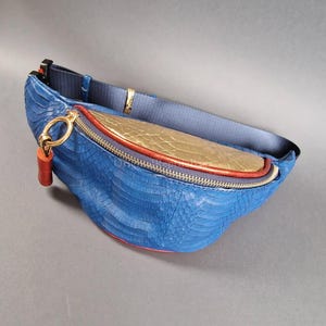 Könnte beinhalten: Blaue Bauchtasche mit Schlangenlederstruktur, mit goldenen und roten Akzenten. Die Tasche hat einen goldenen Reißverschluss und einen roten Lederanhänger. Der Riemen ist marineblau.