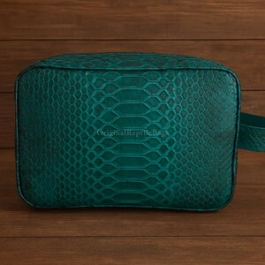 May include: A rectangular, teal-colored bag with a textured, snake-skin pattern. The bag has rounded corners and a matching strap. The text "OriginalReptileBags" is visible on the bag. The bag is set against a wooden background.