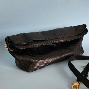 May include: Black clutch with a textured snakeskin pattern. The clutch has a fold-over flap and a detachable black strap with gold-colored clasps. The text "Original Reptile Bags" is visible on the bag.