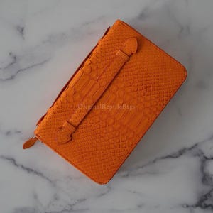 May include: Vibrant orange clutch bag with a reptile skin texture. It has a strap with a clasp and a zip closure. The text "Original Reptile Bags" is visible on the surface.