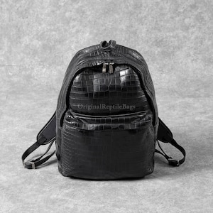 Könnte beinhalten: Schwarzer Rucksack mit strukturierter Krokodilmusterung. Er hat eine Vordertasche, zwei Reißverschlüsse und verstellbare Träger. Der Text "Original Reptile Bags" ist auf der Vorderseite aufgedruckt.