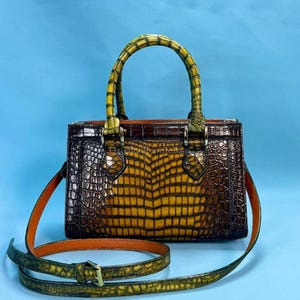 May include: A brown and yellow crocodile-textured handbag with a top handle and a detachable shoulder strap. The bag has a rectangular shape with a structured design. The handle and strap are green and the interior is orange.