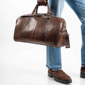 Könnte beinhalten: Eine braune Leder-Reisetasche mit Reptilienhautmuster. Die Tasche hat einen oberen Griff, einen Schultergurt und einen Reißverschluss. Der Text "Original Reptile Bags" ist auf der Tasche aufgedruckt.