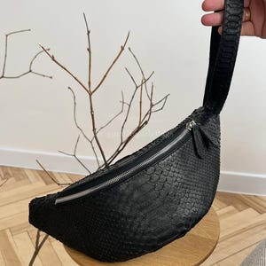 Könnte beinhalten: Schwarze Bauchtasche mit einem strukturierten, schuppenartigen Muster. Die Tasche hat eine gebogene Form, einen silbernen Reißverschluss und einen passenden Riemen. Die Tasche steht auf einem kleinen Holzschemel.