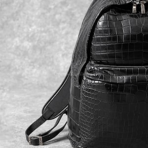Puede incluir: Una mochila negra con un patrón texturizado de piel de cocodrilo. La bolsa incluye un compartimento principal con cierre de cremallera, un bolsillo frontal y correas ajustables para los hombros con hebillas metálicas.