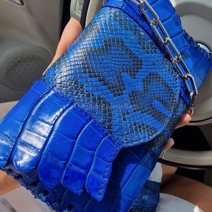 Könnte beinhalten: Eine leuchtend blaue Clutch mit silberner Kette. Die Tasche hat ein strukturiertes, krokodilartiges Muster auf der Klappe und ein kontrastierendes Schlangenhautdesign auf dem Körper. Ein auffälliges, helles Accessoire.
