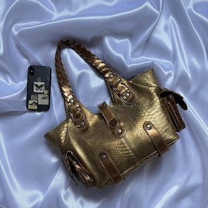 Könnte beinhalten: Eine goldfarbene Handtasche mit Schlangenlederstruktur. Die Tasche hat einen geflochtenen Griff und mehrere Taschen. Die Tasche liegt auf einer weißen Satin-Oberfläche. Ein Mobiltelefon mit Schmetterlingsdesign liegt neben der Tasche.