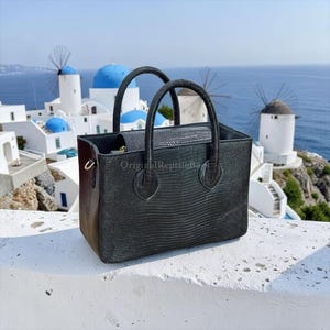 Può includere: Borsa tote nera con due manici, caratterizzata da un motivo strutturato simile a quello di un rettile. La borsa ha una forma rettangolare e una pochette più piccola abbinata all'interno. Lo sfondo mostra una vista panoramica di edifici bianchi, cupole blu e mulini a vento.