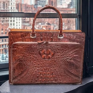 May include: A brown leather briefcase and matching handbag set with a crocodile skin texture. The briefcase has a zip closure and the handbag has a top handle. The set is displayed in front of a window with a city view.