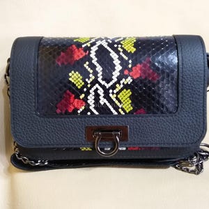 Puede incluir: Un bolso bandolera gris oscuro con un panel de piel de serpiente con detalles rojos, amarillos y blancos. El bolso tiene forma rectangular, un cierre metálico y una correa de cadena plateada. La piel tiene una apariencia texturizada.