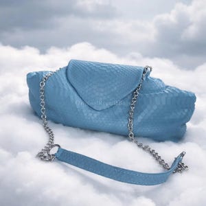 Könnte beinhalten: Hellblaue Clutch-Tasche mit Klappenverschluss und silberner Kette. Die Tasche besteht aus einem strukturierten Material, möglicherweise Leder oder lederähnlichem Material. Der Text "Original Reptile Bags" ist sichtbar.