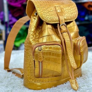 Könnte beinhalten: Ein metallisch goldener Rucksack mit einer strukturierten Krokodilleder-Optik. Die Tasche verfügt über eine obere Klappe mit Schnallenverschluss, zwei vordere Außentaschen mit vertikalen Reißverschlüssen und verstellbare Schultergurte mit hellbraunen Zierleisten.