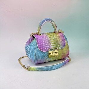 Puede incluir: Un pequeño bolso colorido con un patrón de piel de serpiente arcoíris. El bolso tiene un cierre dorado, una correa de cadena dorada y un asa blanca. Los colores incluyen azul, morado, amarillo y verde.